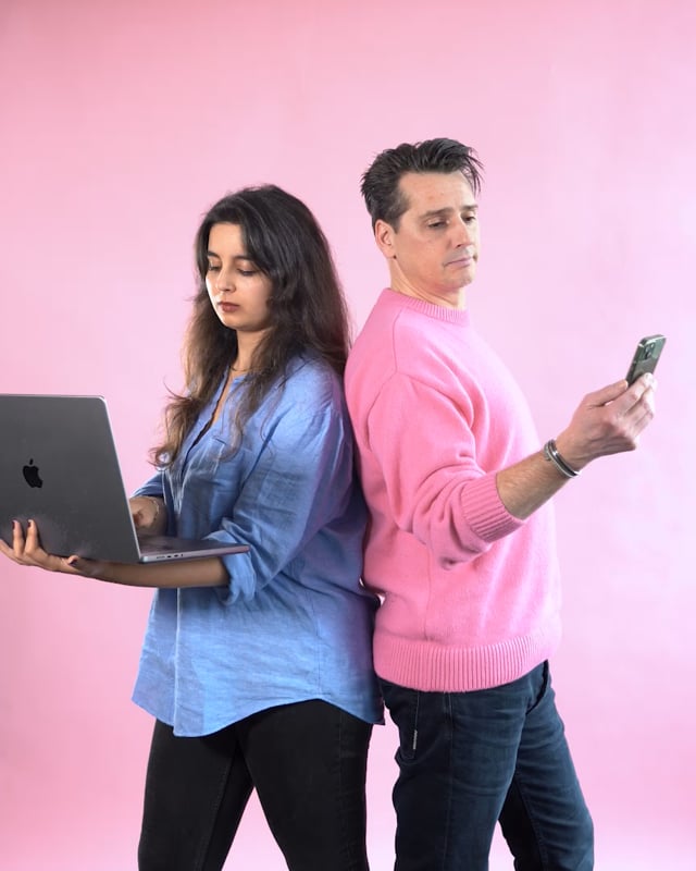 Een vrouw in een blauwe blouse houdt een laptop vast en kijkt gefocust naar het scherm, terwijl een man in een roze trui met zijn rug tegen haar aan leunt en aandachtig op zijn telefoon kijkt. De achtergrond is roze.